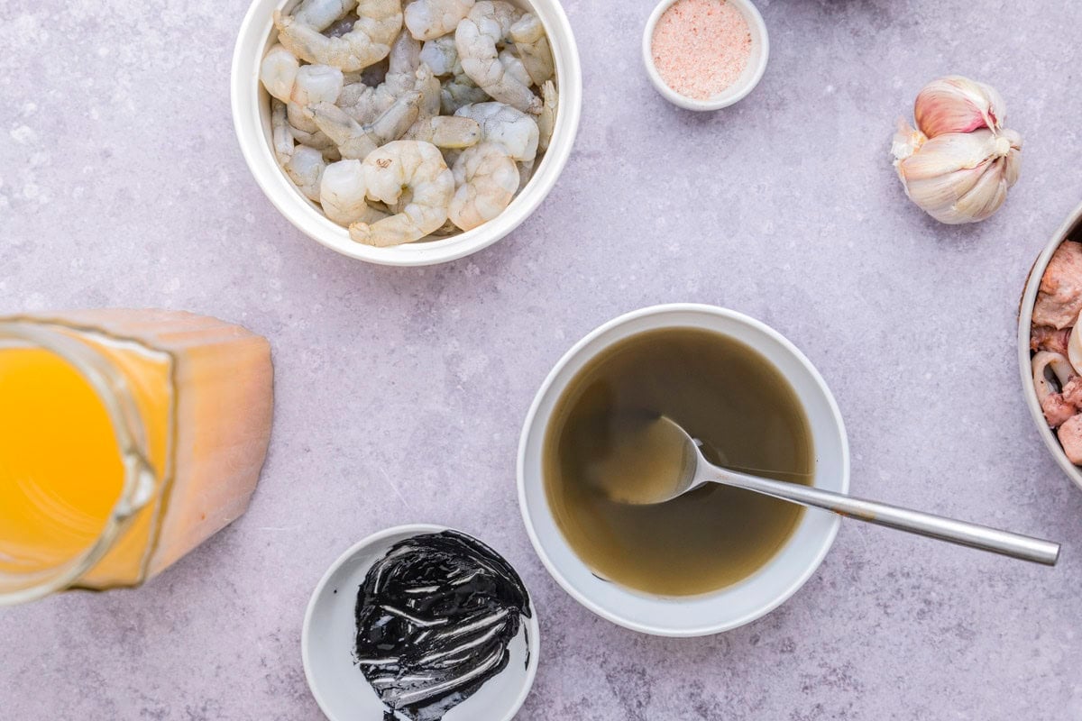 bowls on counter filled with fish stock, squid ink, and raw shrimp.