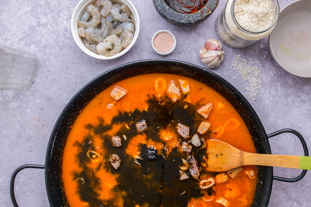 black squid ink being mixed into red paella ingredients in large flat black pan.