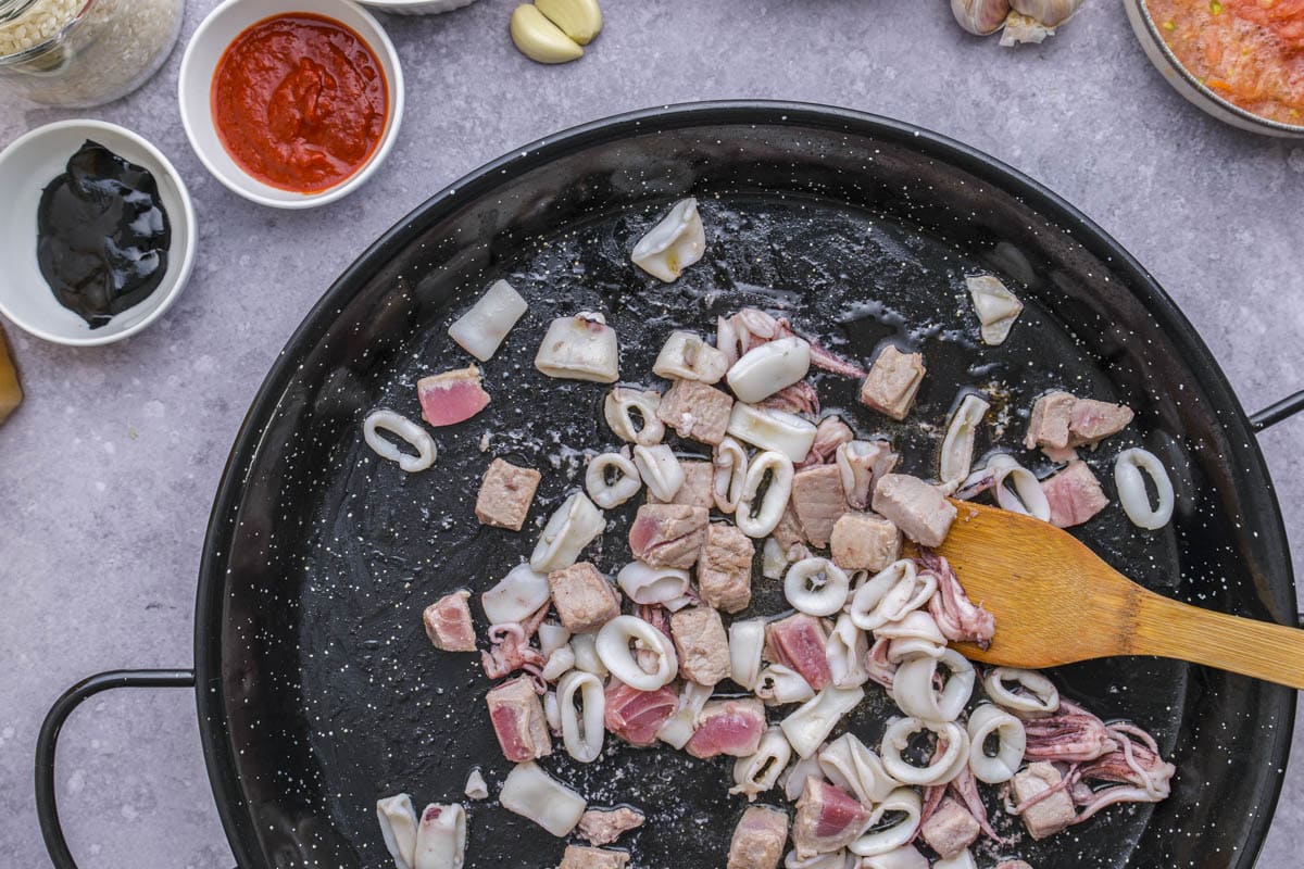 pieces of seafood frying in large black pan being stirred with wooden spatula.