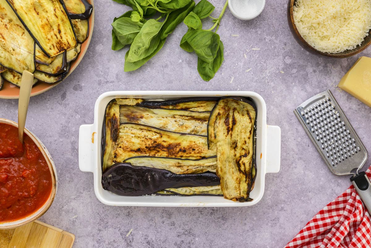 layer of grilled eggplant in white baking dish on counter with ingredients around.
