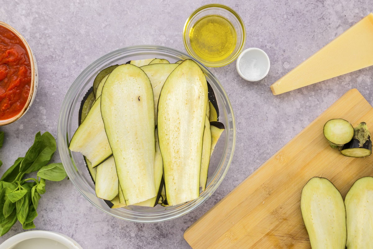 slices of raw eggplant in bowl with counter top and ingredients around.