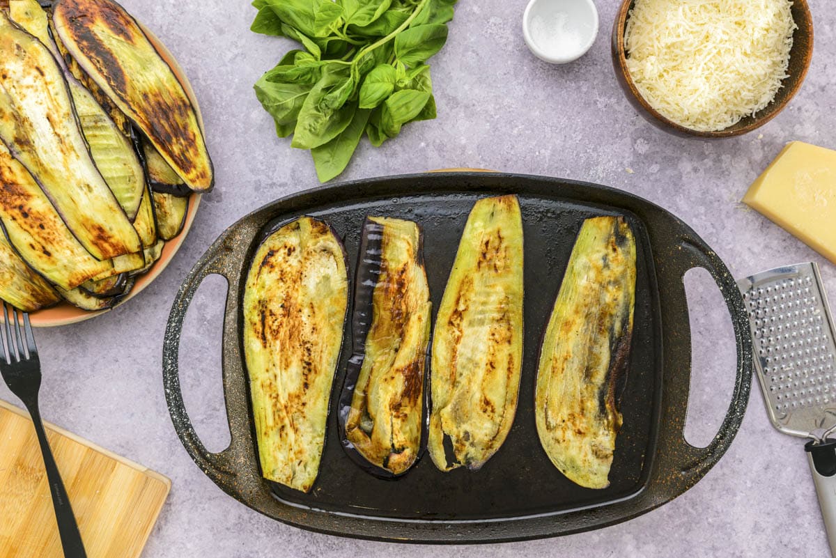 small grill frying slices of eggplant with other ingredients around on counter.