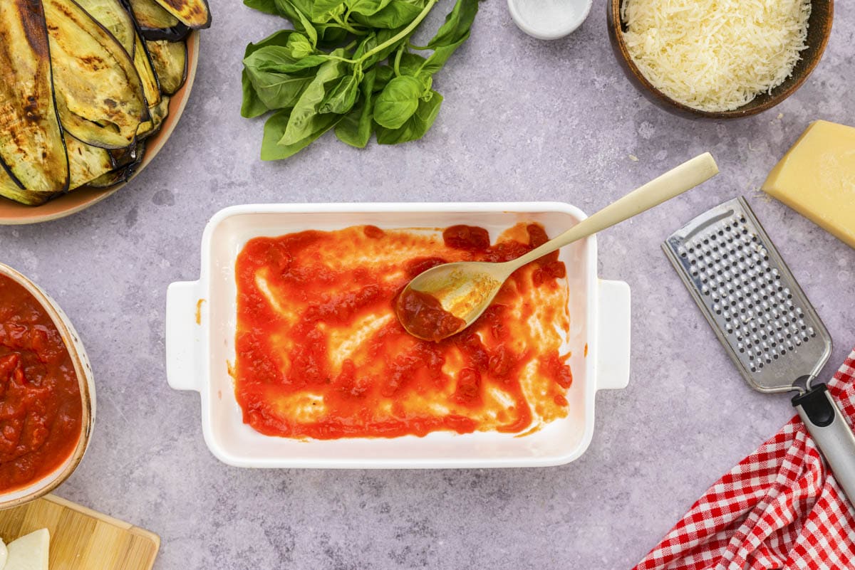 wooden spoon spreading tomato sauce into white baking dish.