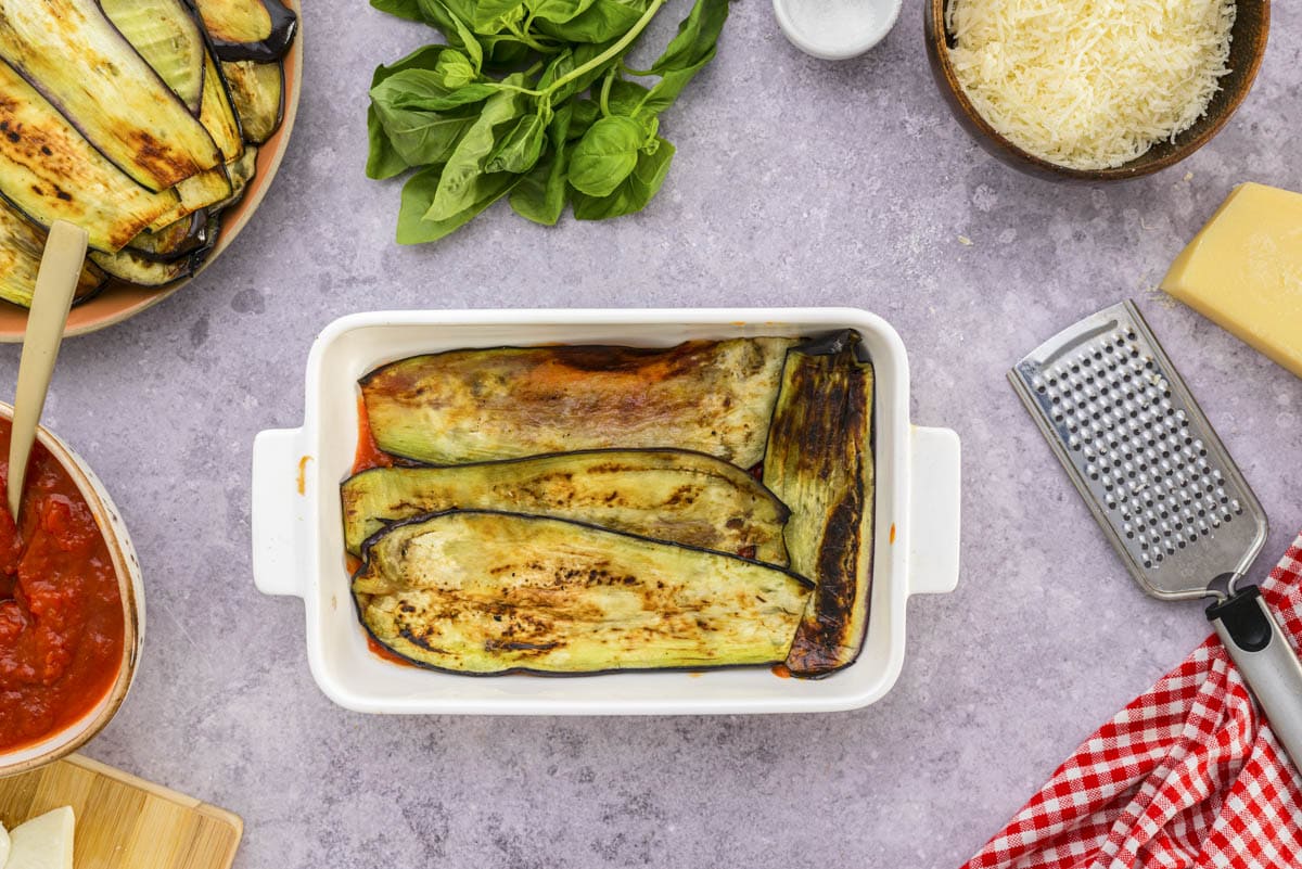 layer of grilled eggplant laid in white baking dish on counter.