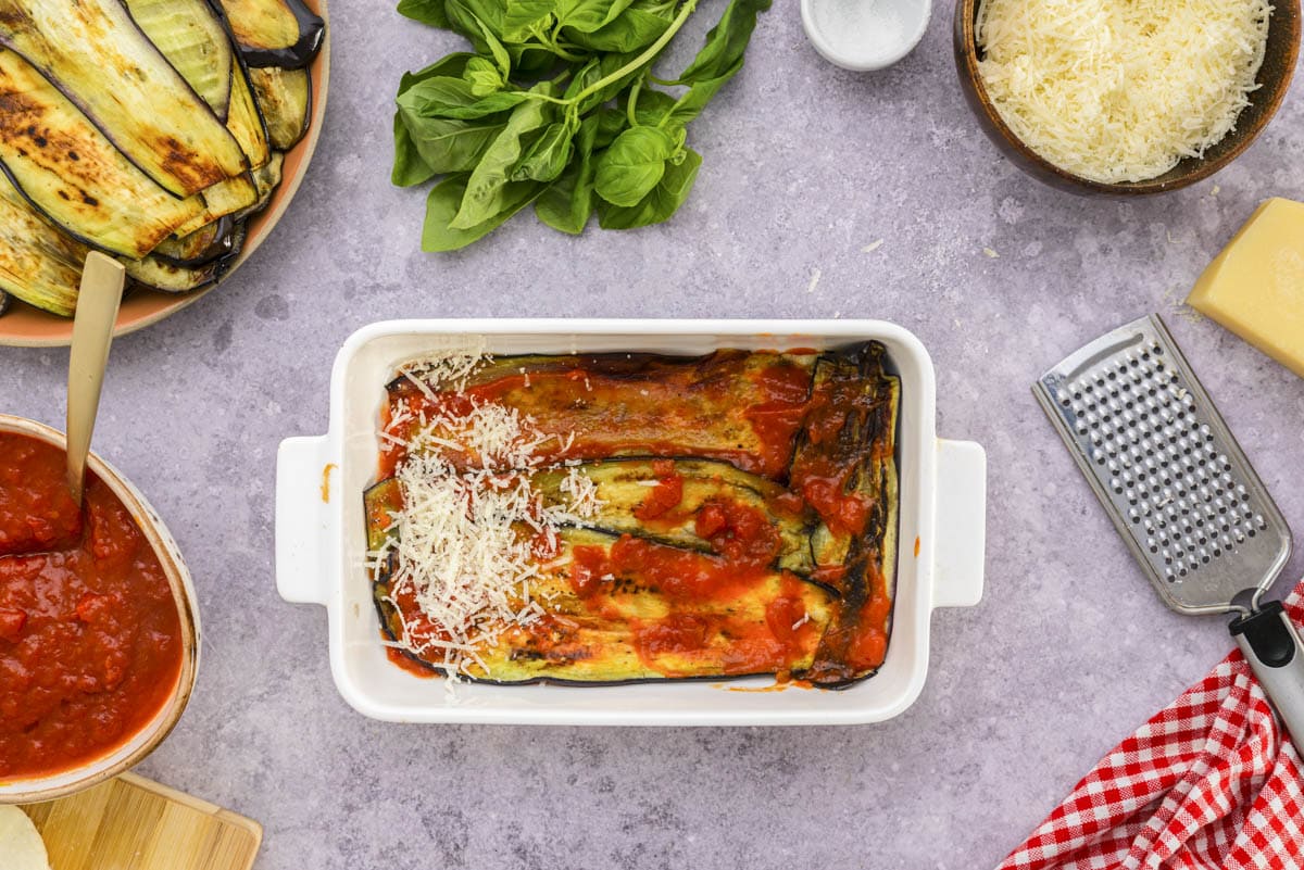 grated parmesan on layers of tomato sauce and eggplant in white baking dish.