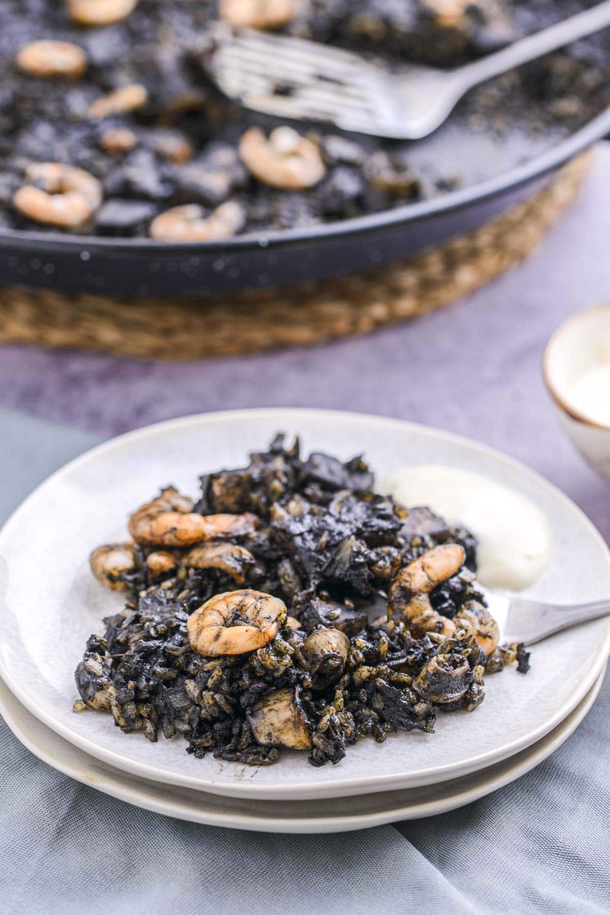 plate of squid ink paella with silver fork sticking out and larger pan behind.