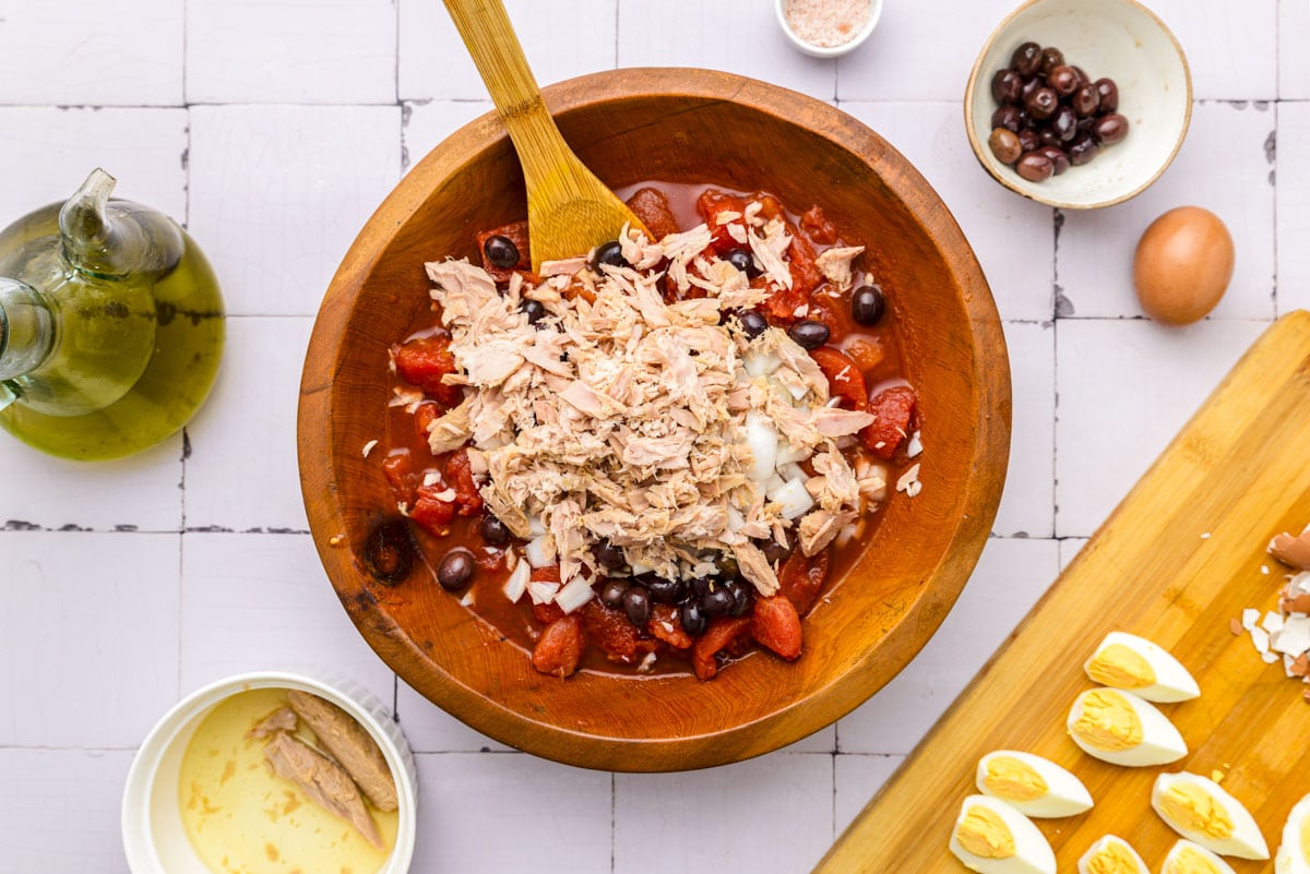 wooden spoon sticking out of wooden bowl of tomatoes, tuna, onions, and more.