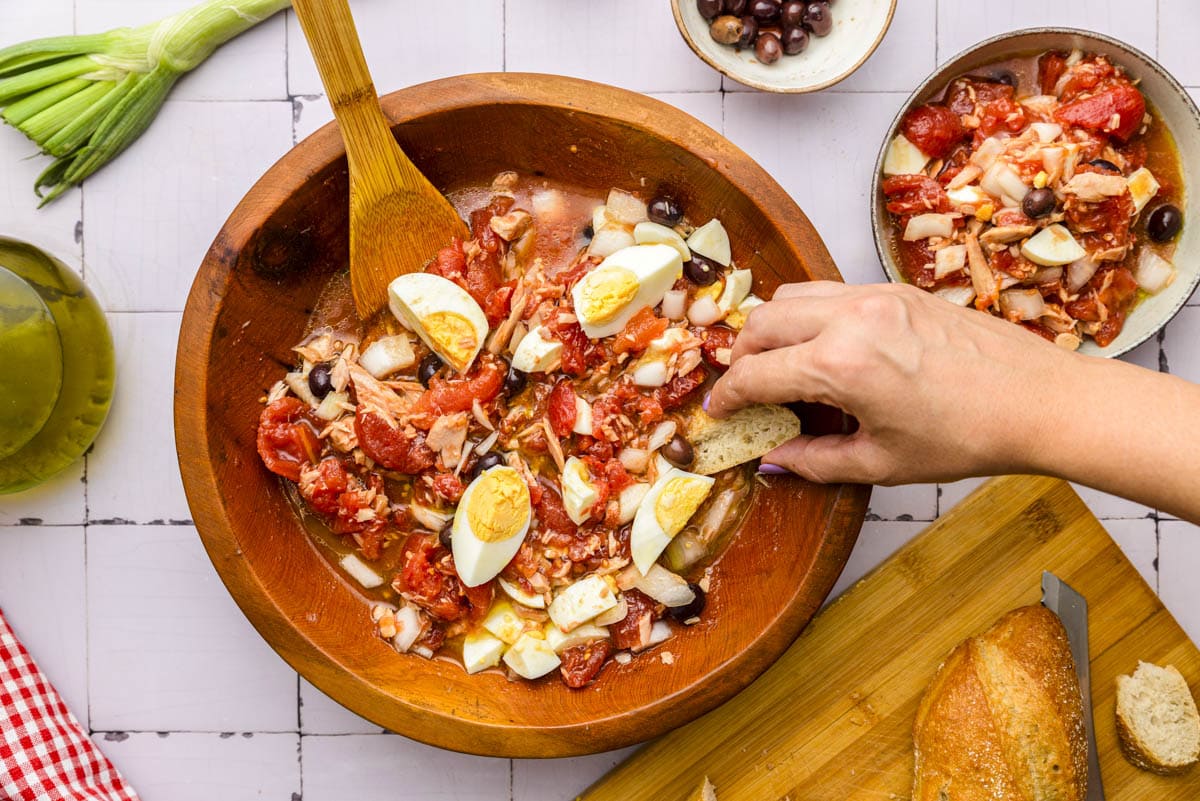 hand dipping bread into juicy tomato salad in wooden bowl on counter.