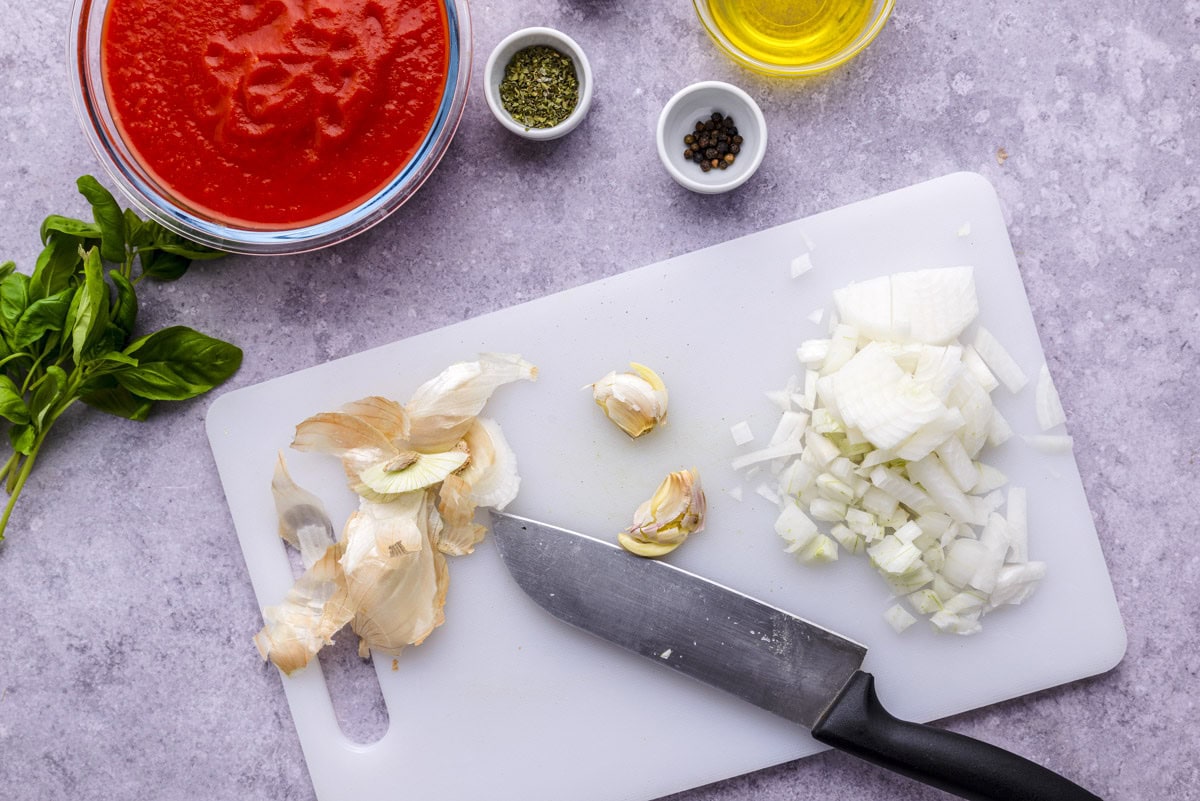 garlic cloves and diced onions on white cutting board with knife beside.