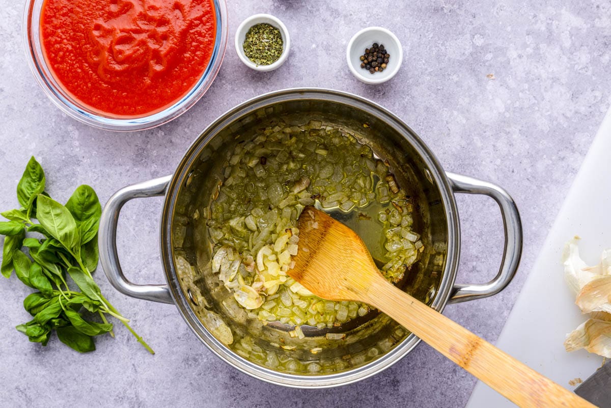 wooden spoon stirring onions and garlic in olive oil in silver pan.