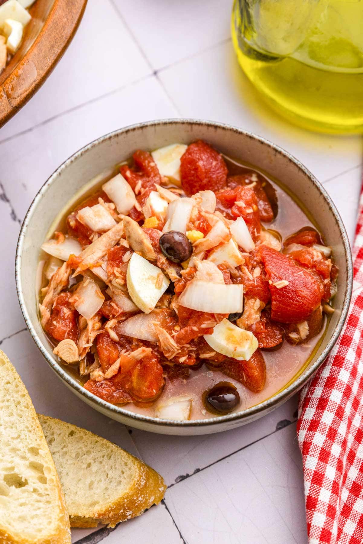 bowl of tomatoes, olives, and boiled egg sitting on counter with sliced bread around.