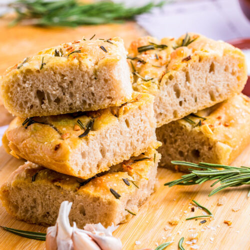 slices of focaccia stacked on wooden board with rosemary beside.
