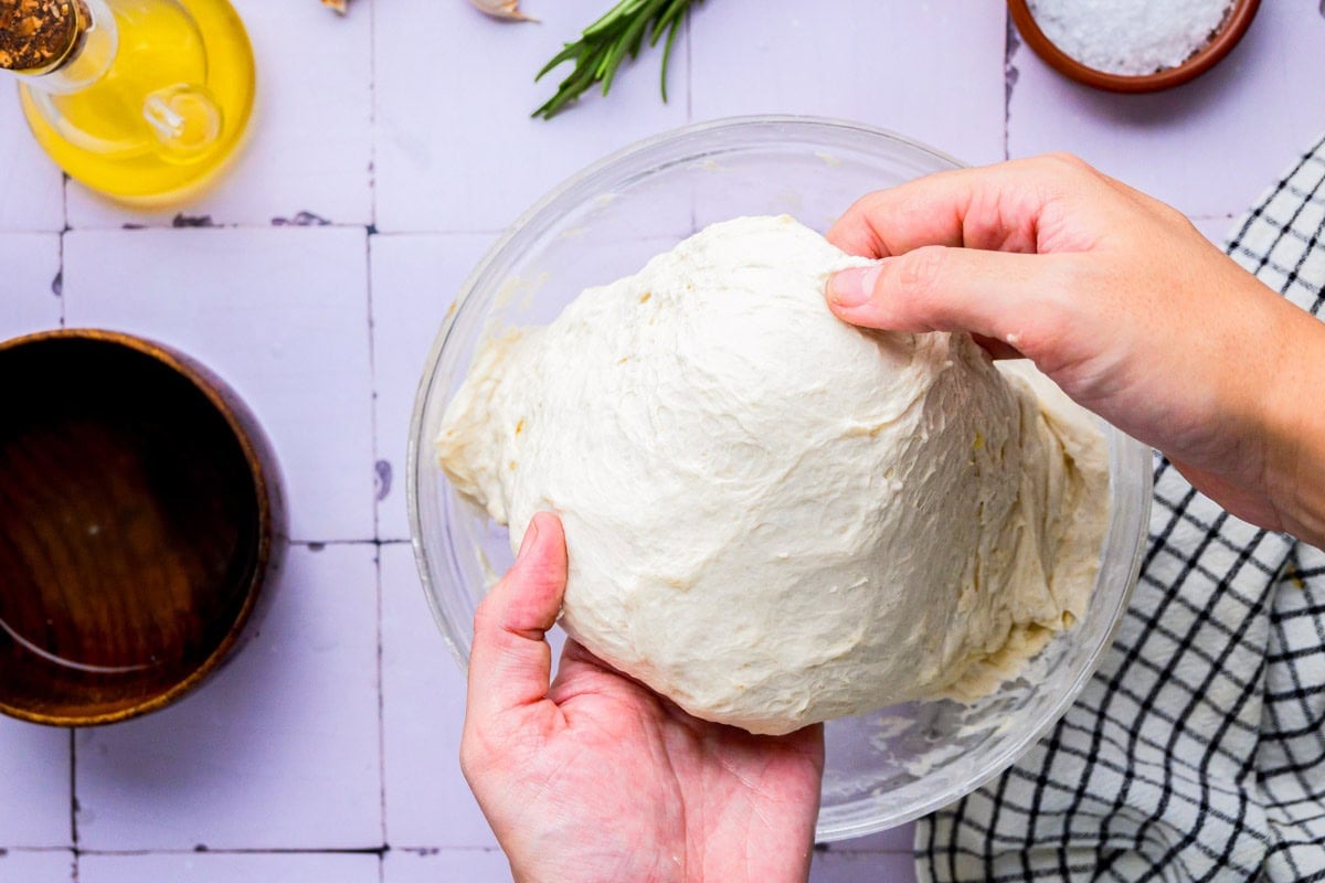 hands stretching dough above clear glass bowl with counter around.