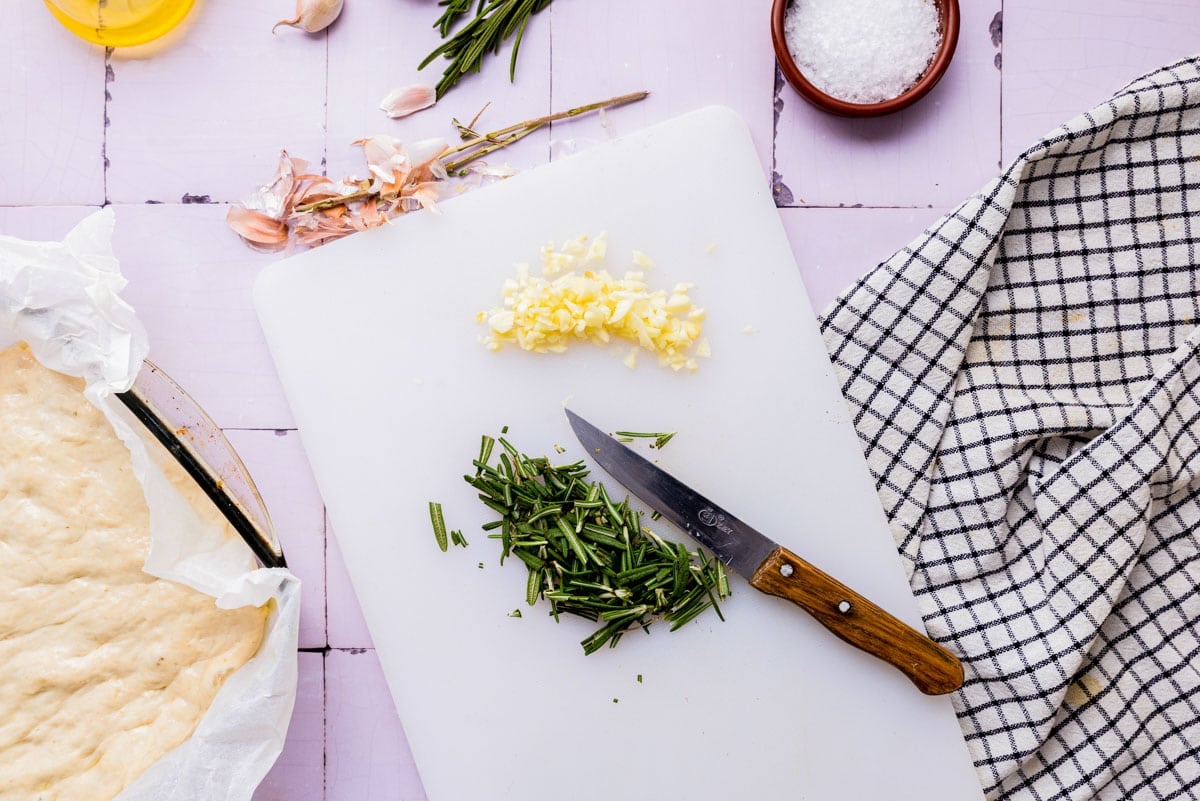 knife on cutting board with rosemary chopped up beside chopped garlic cloves.