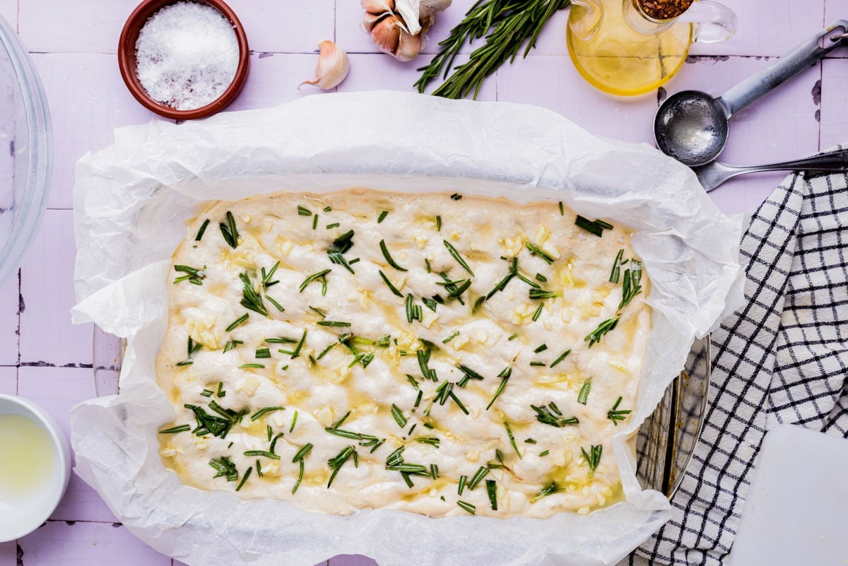 chopped rosemary on top of raw focaccia dough in pan.