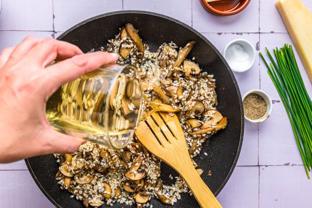 hand pouring white wine into pan of rice and mushrooms.