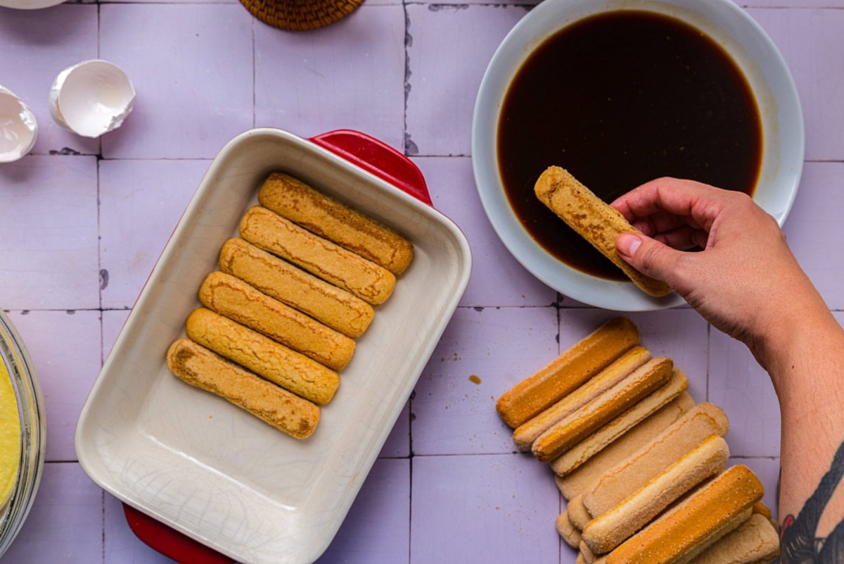 hand dipping ladyfinger into coffee and laying them into dish.