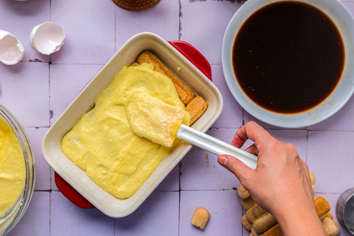 hand spreading sweet cheese mixture into ladyfingers in baking pan.
