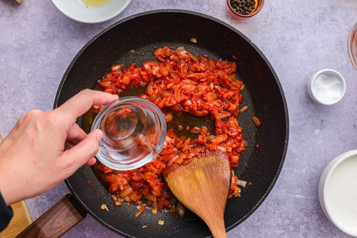 hand pouring vodka into red sauce in frying pan.