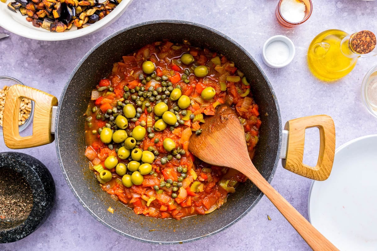 capers and green olived added to black frying pan of cooked vegetables.