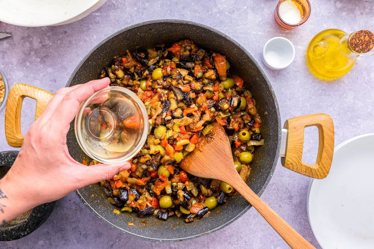 hand adding clear liquid to frying pan of cooked vegetables with wooden spoon.