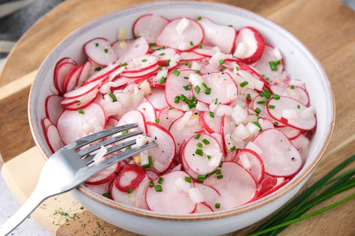 silver fork piercing sliced radish salad in bowl.
