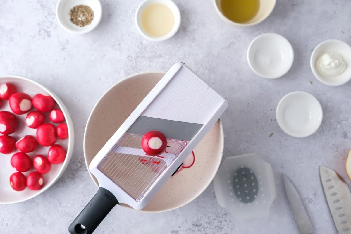 radish being sliced on a mandolin slicer above a bowl.