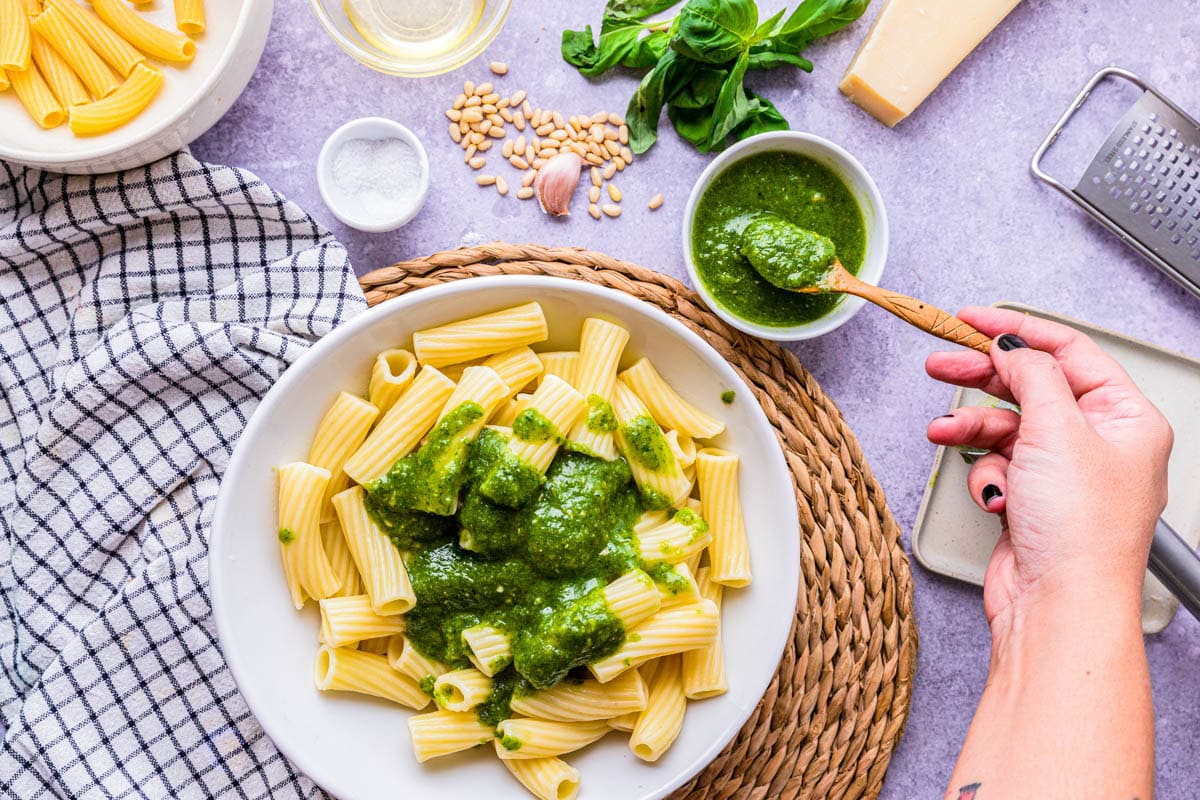 hand spooning green pesto sauce onto cooked noodles in bowl.