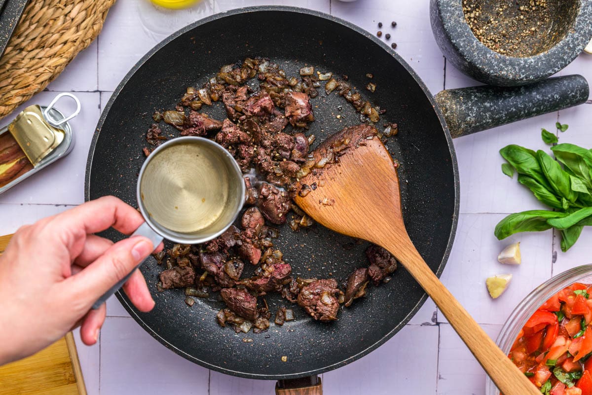 cooked chicken livers in frying pan with white wine being added via measuring cup.