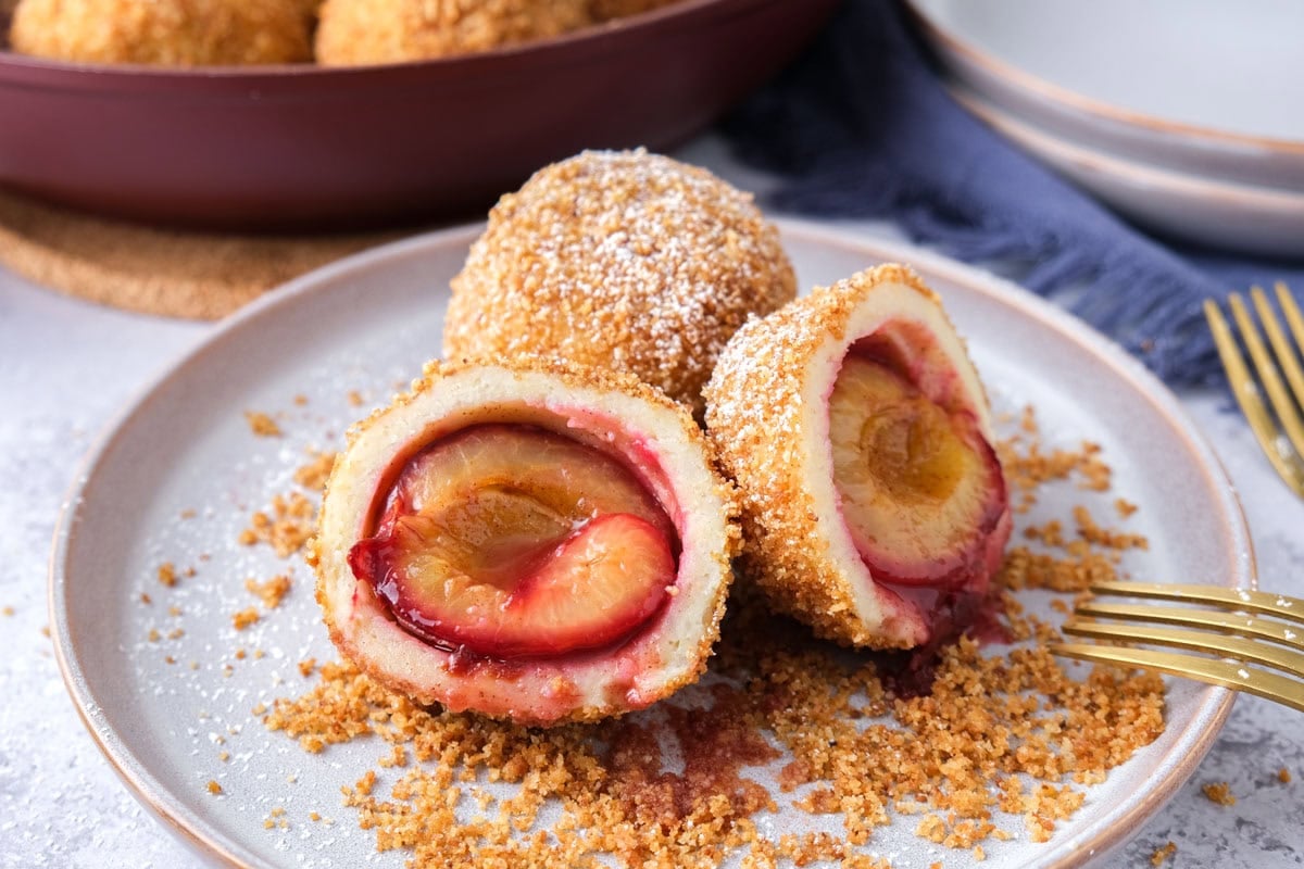 plum dumplings cut open on plate with forks and breadcrumbs around.