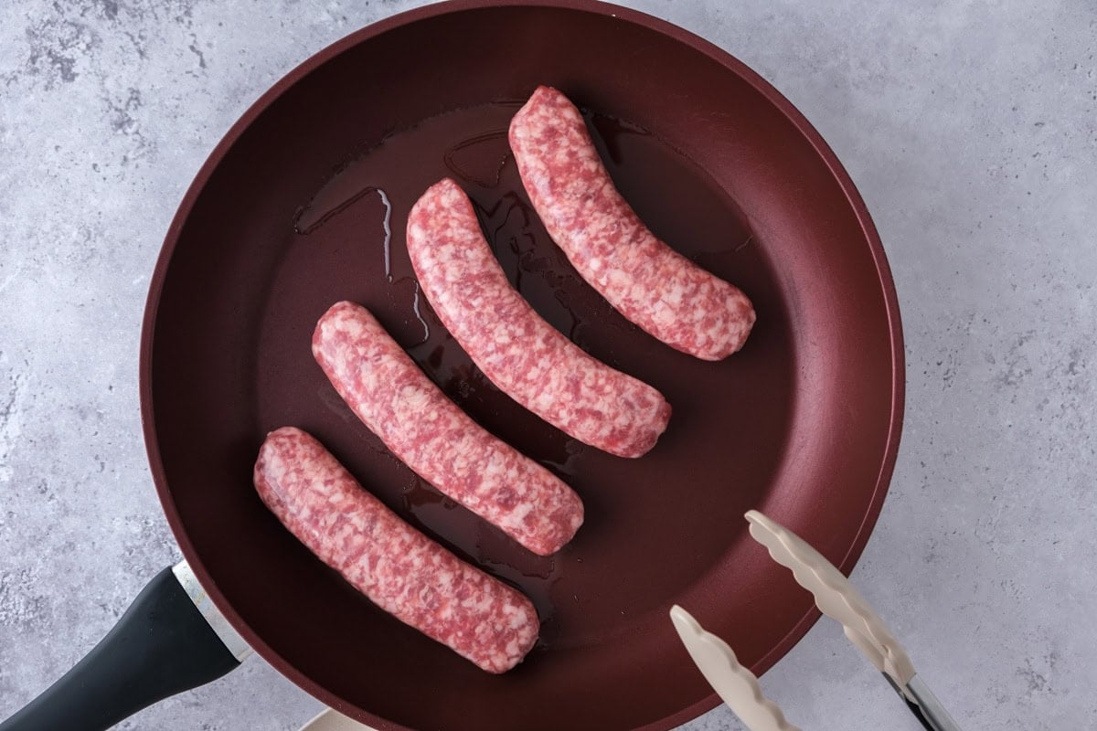 four raw bratwurst sausages in frying pan with tongs beside.
