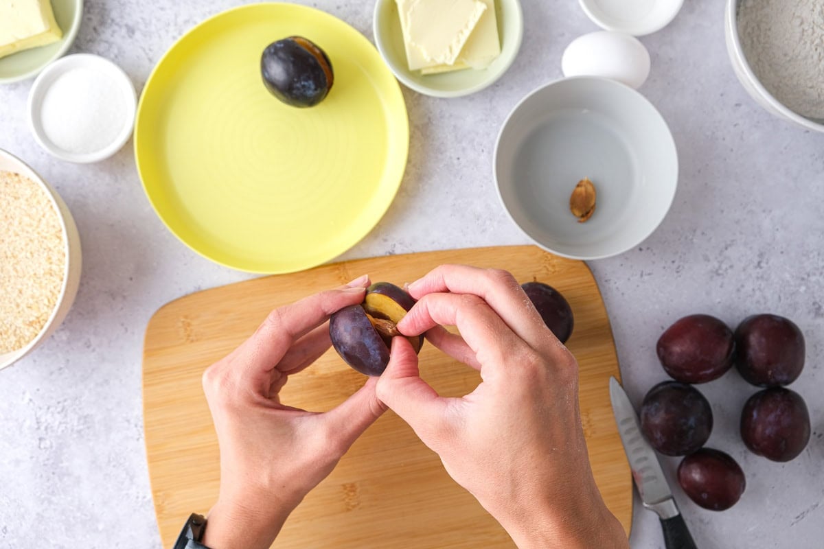 hand removing a pit from a plum with ingredients below on counter.