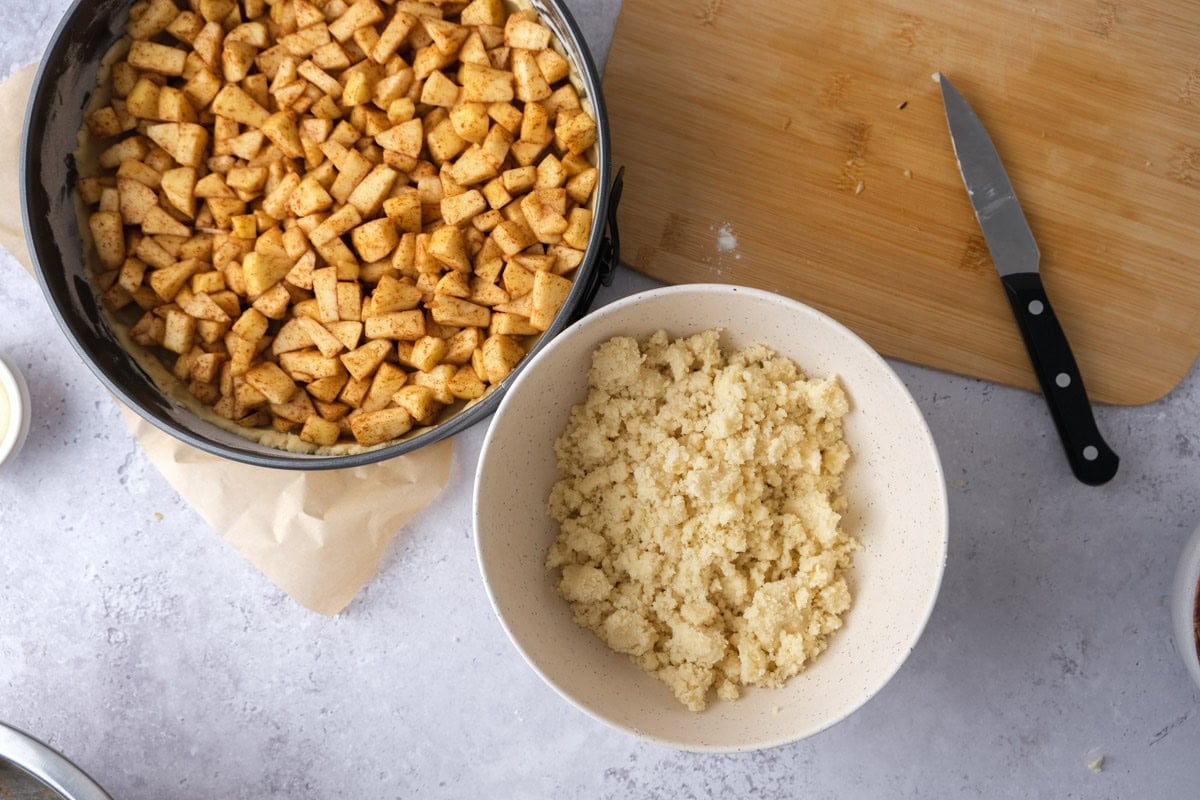 dough to make cake crumbles in bowl beside raw apple cake in pan.